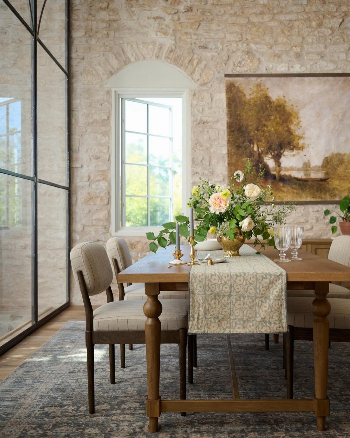 Wooden dining table set with floral centerpiece and runner against a stone wall.