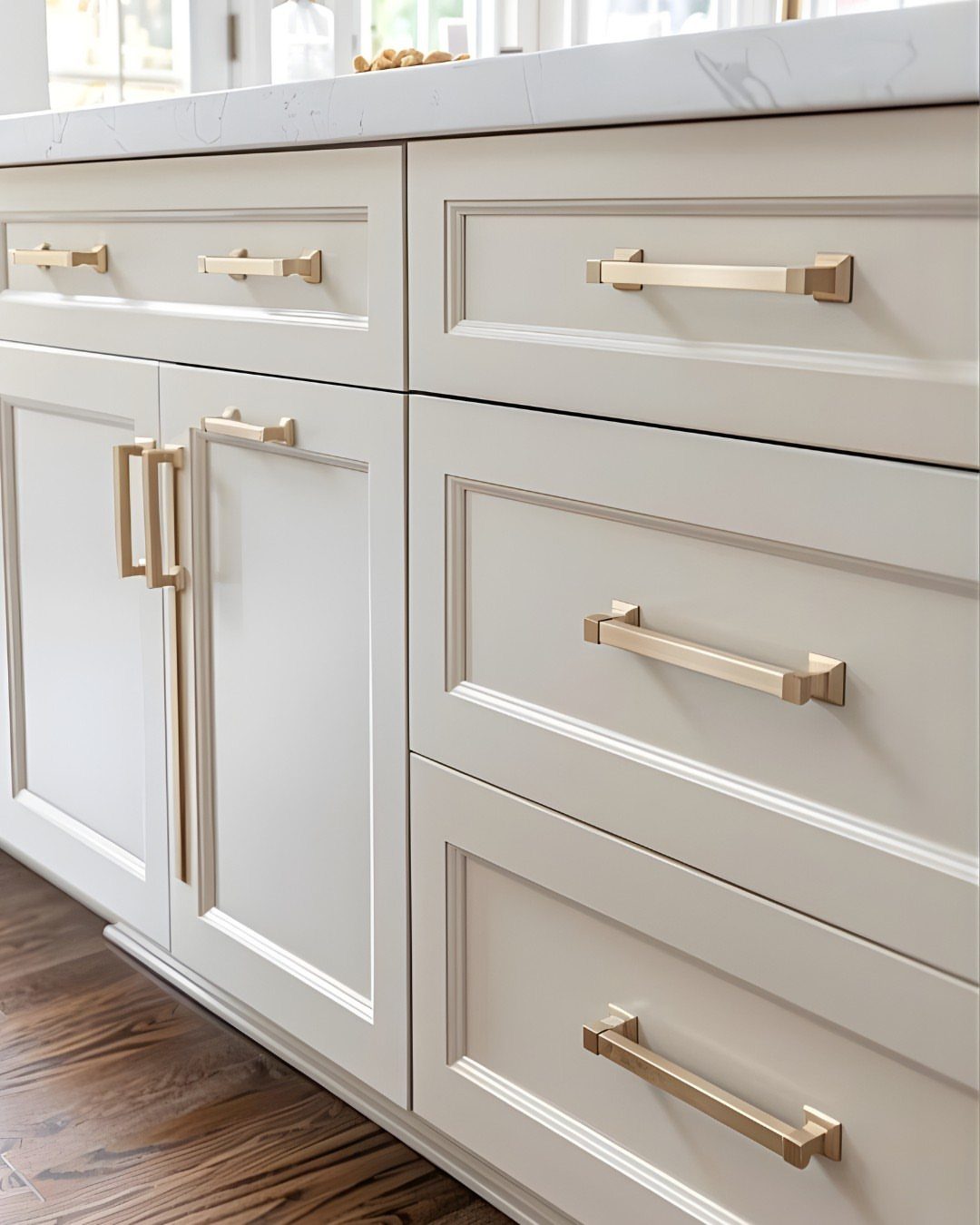 Off-white kitchen cabinets and drawers with sleek gold hardware and marble countertop.