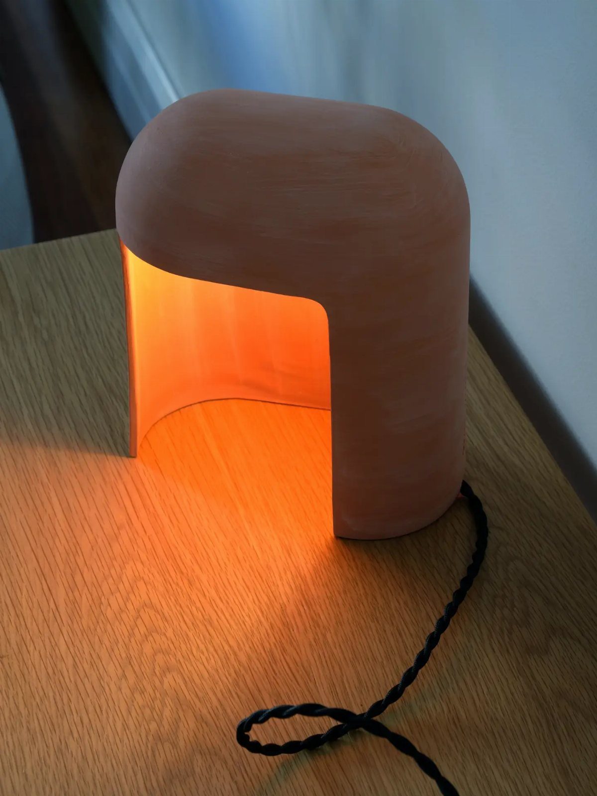 Terracotta ceramic table lamp with a curved design emitting warm light on wood.