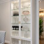 White glass-door display cabinet with interior lighting showcasing crystal glassware and bowls.