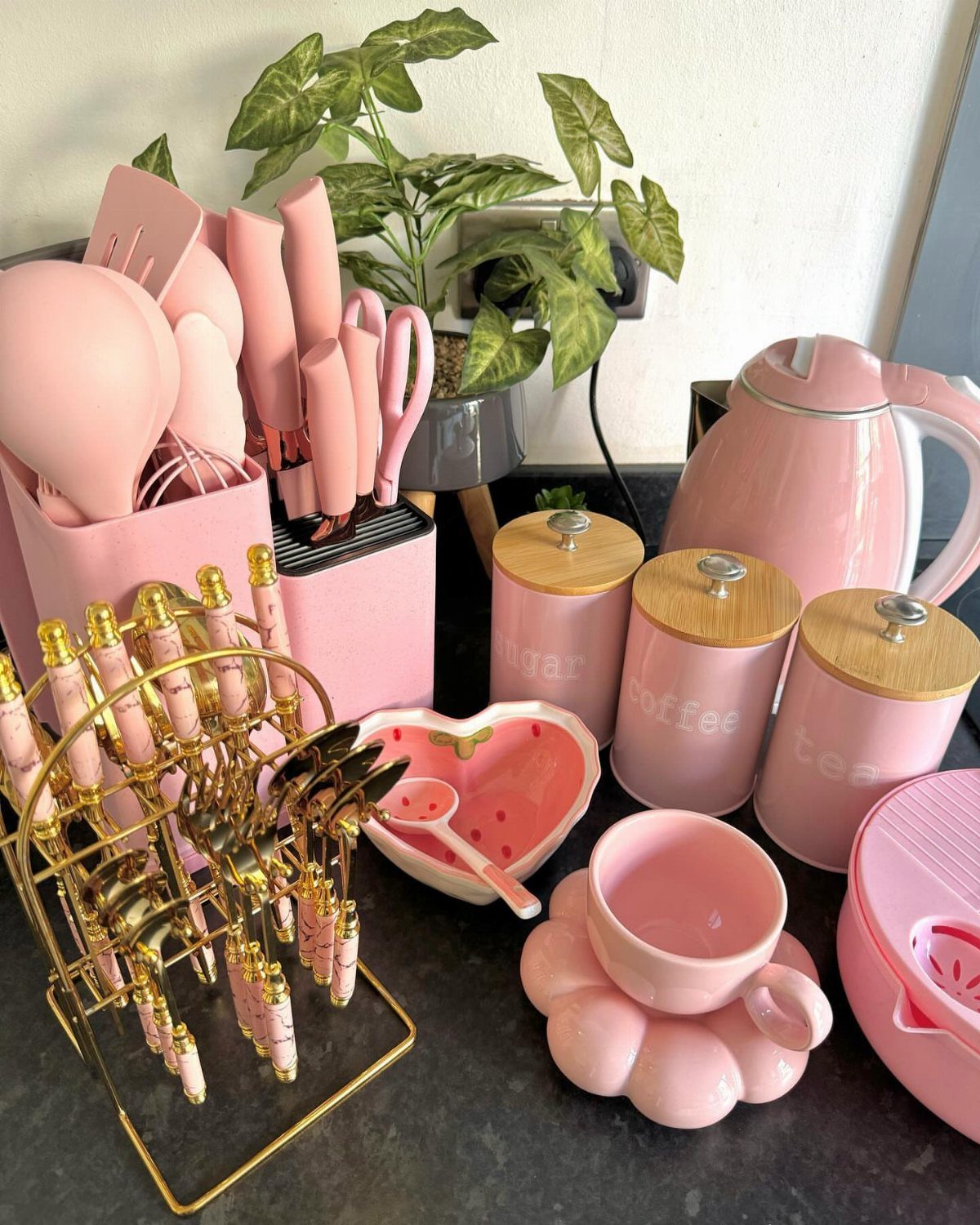 Pink kitchen accessories including a kettle, canisters, and utensils on a dark countertop.