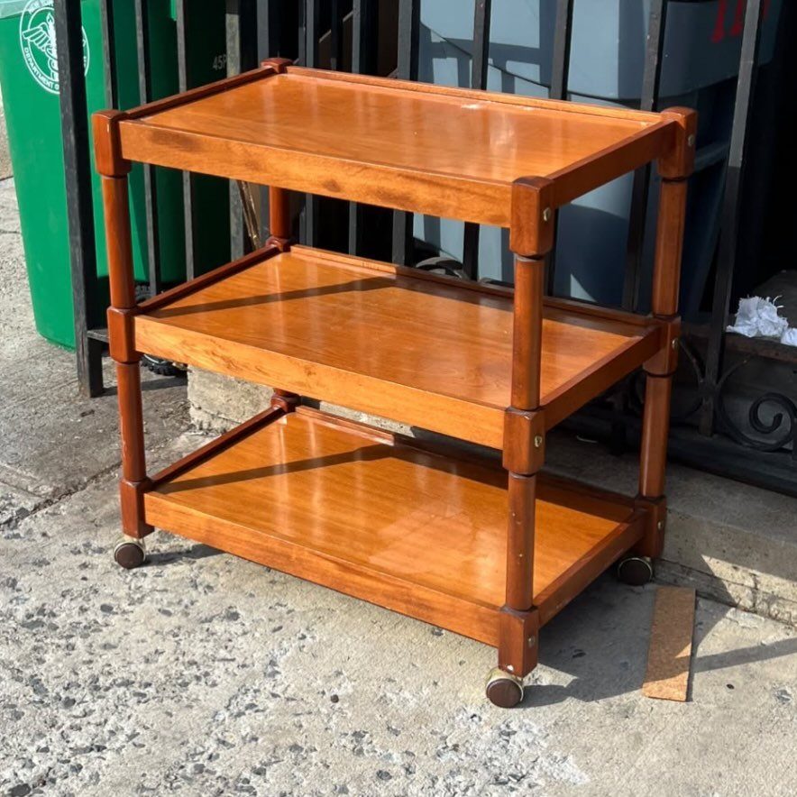 Three-tiered brown wooden shelving unit with small caster wheels on a concrete surface.