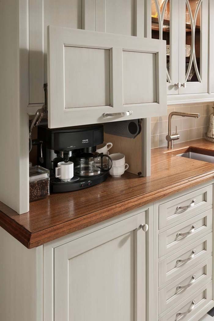 Coffee maker and mugs inside a white cabinet with a lift-up door.