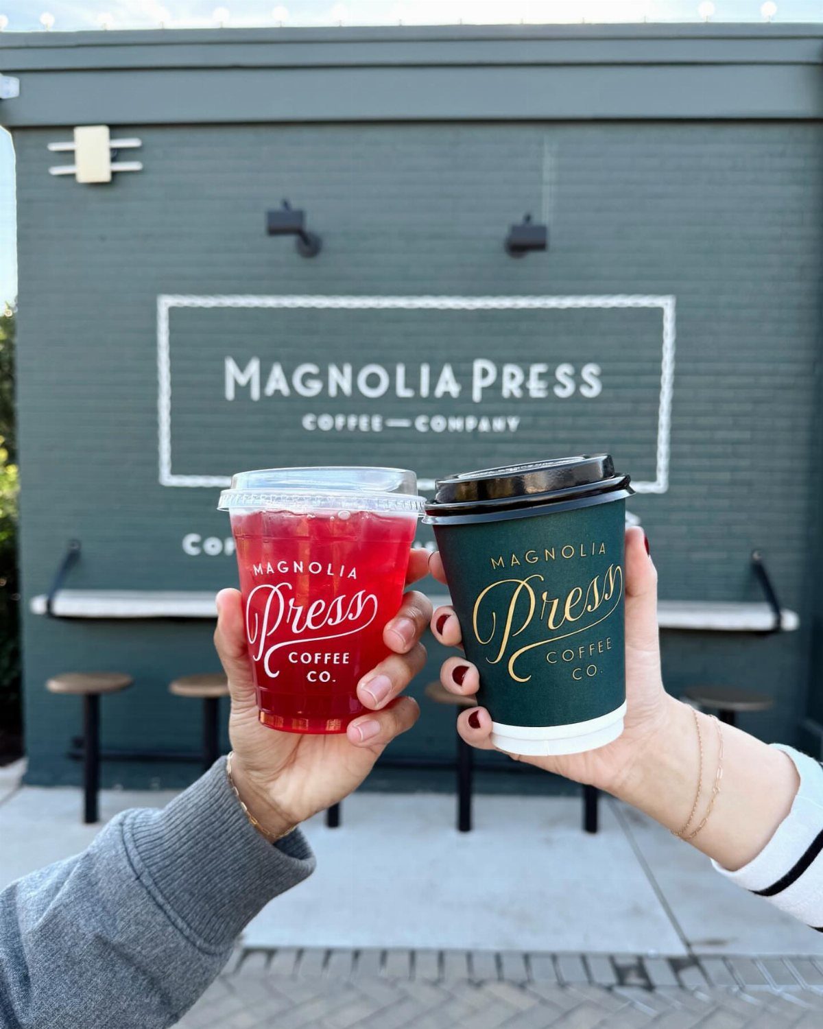 Two coffee drinks held in front of the Magnolia Press Coffee Company storefront.