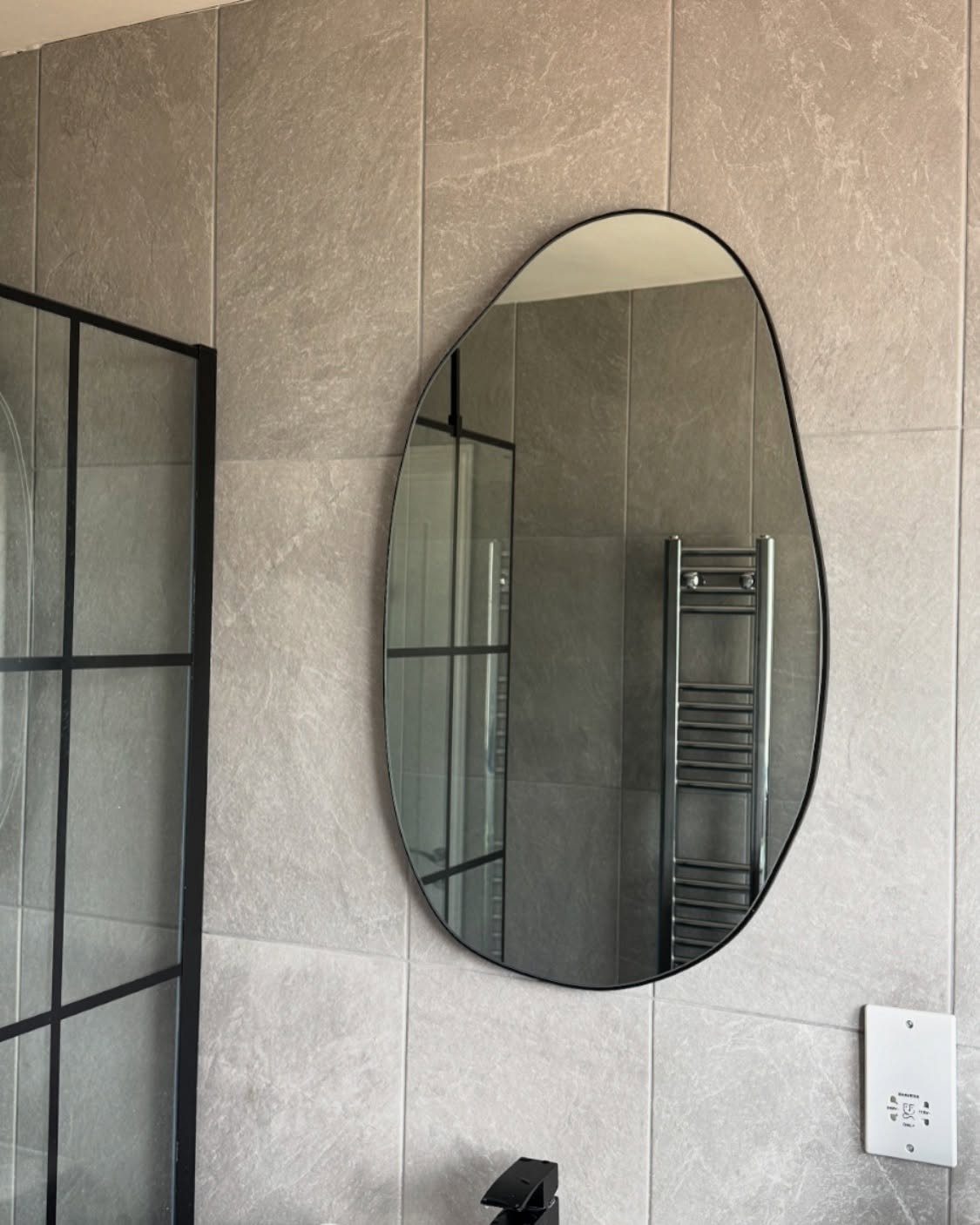 Irregularly shaped organic mirror mounted on a grid of light grey bathroom wall tiles.