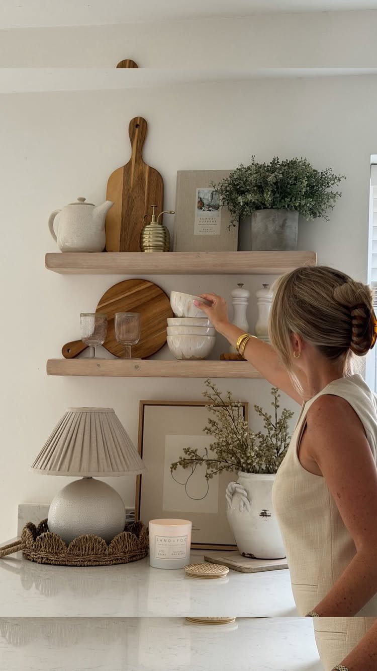 Woman reaching for a white bowl on light wood open kitchen shelves.