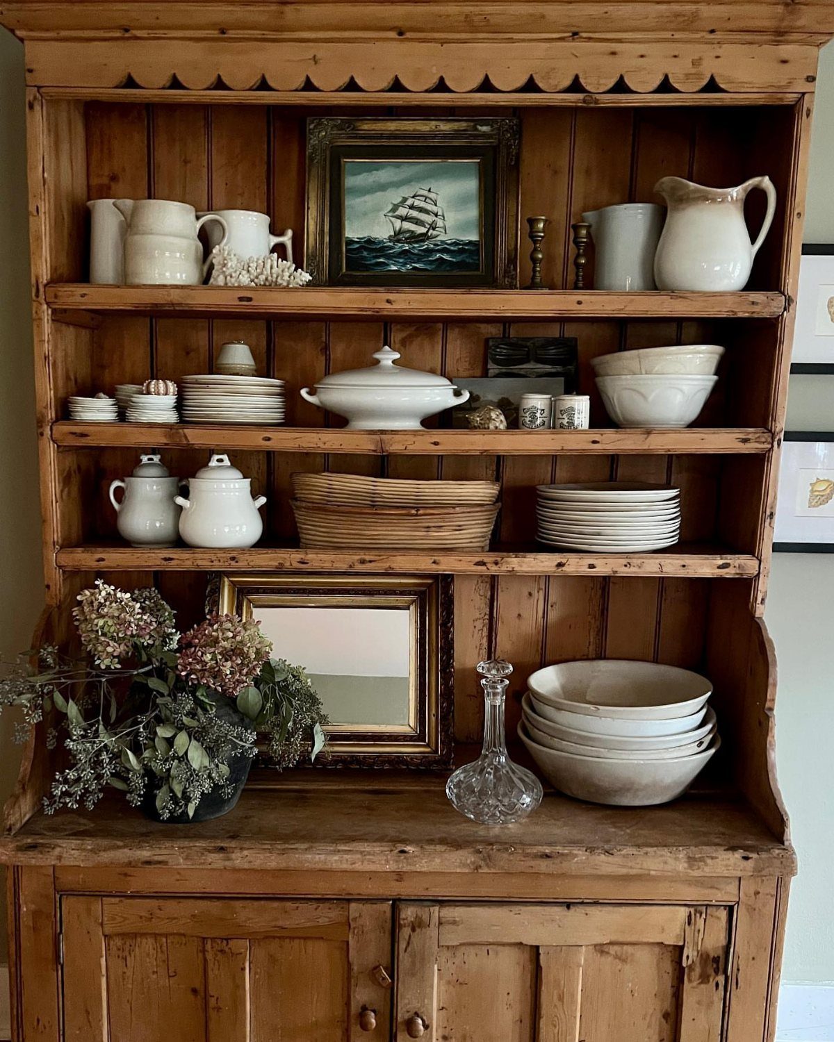 Rustic wooden painter's cabinet with white ceramics, a small painting, and dried flowers.