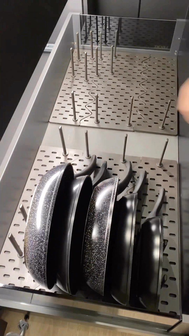 Black frying pans stored vertically in a grey metal pegboard drawer organizer tray.