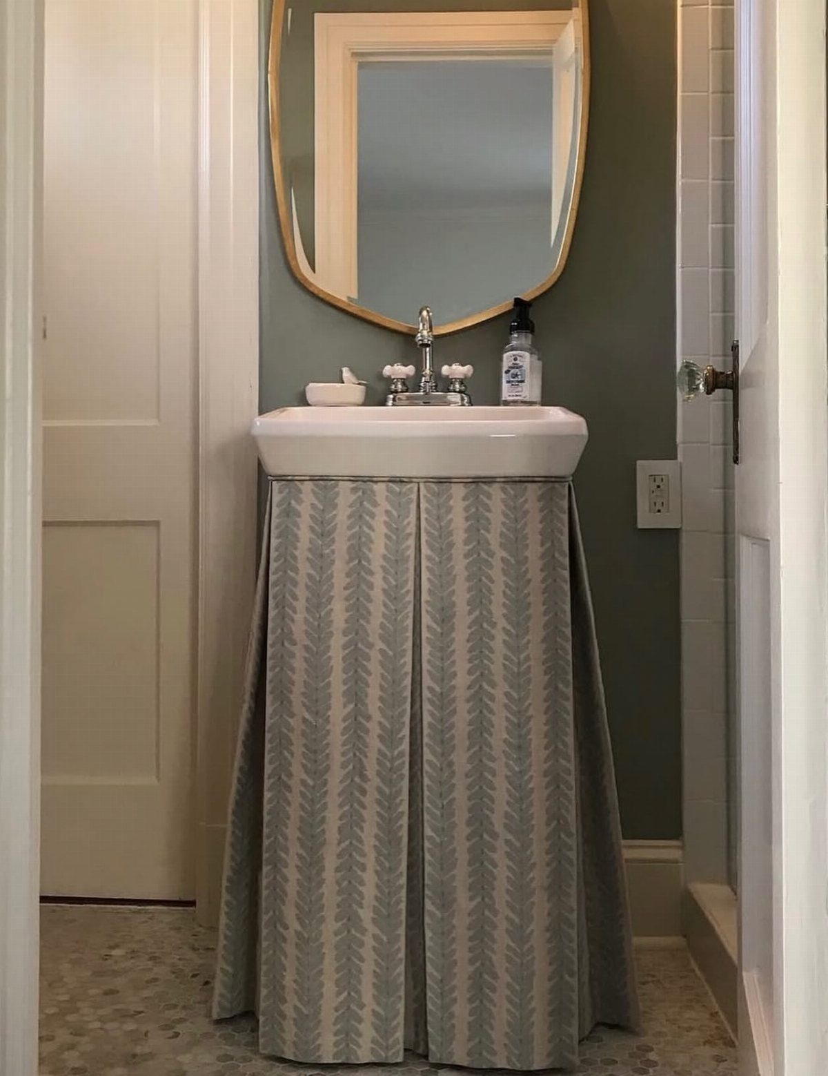 White pedestal sink with a grey and white patterned fabric skirt and gold mirror.
