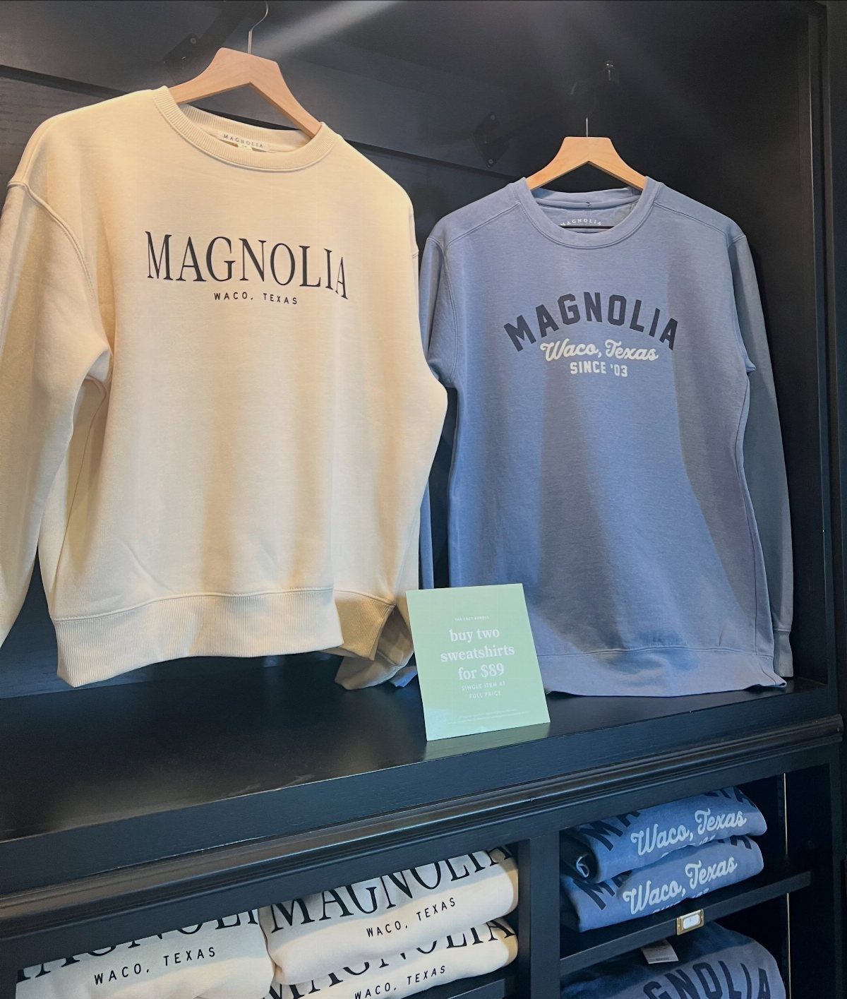 Magnolia branded sweatshirts in cream and blue displayed on a black retail shelf.
