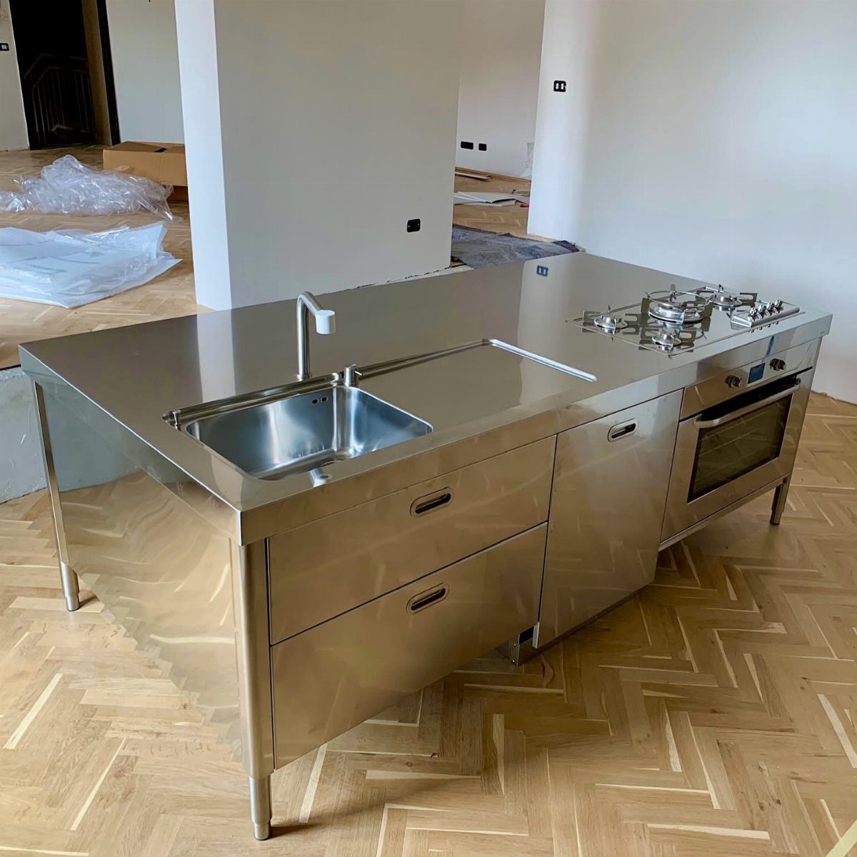 Stainless steel kitchen island with integrated sink and stovetop on herringbone wood flooring.