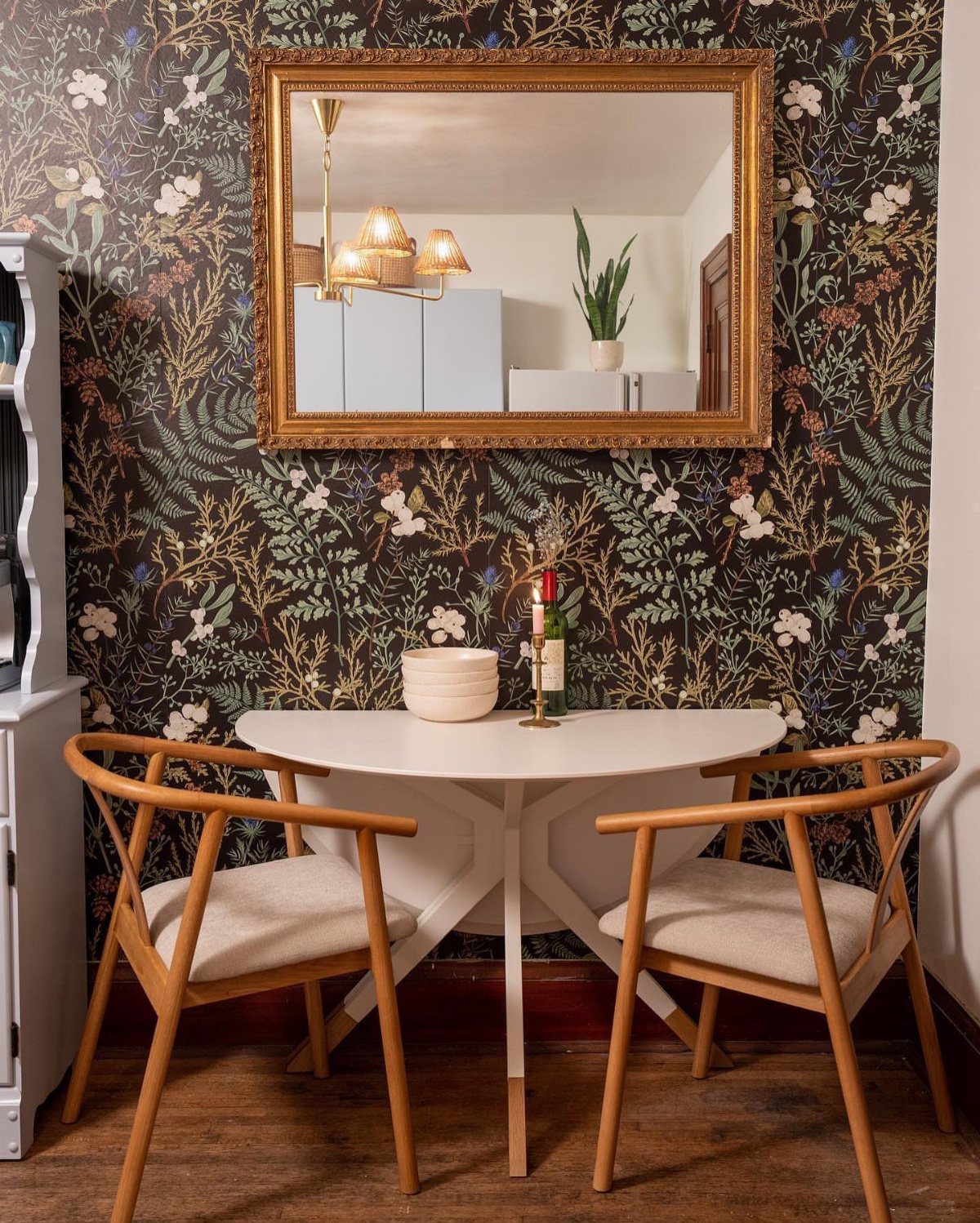 White round dining table pushed against a wall with dark floral wallpaper and mirror.