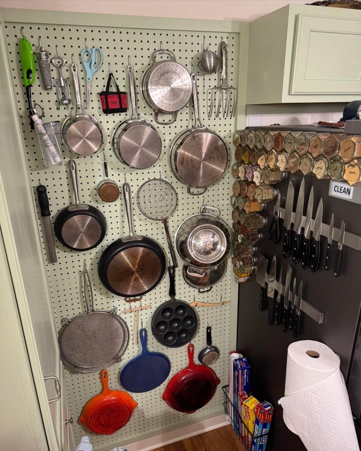 Wall pegboard rack holding various pots, pans, and kitchen utensils in a kitchen.