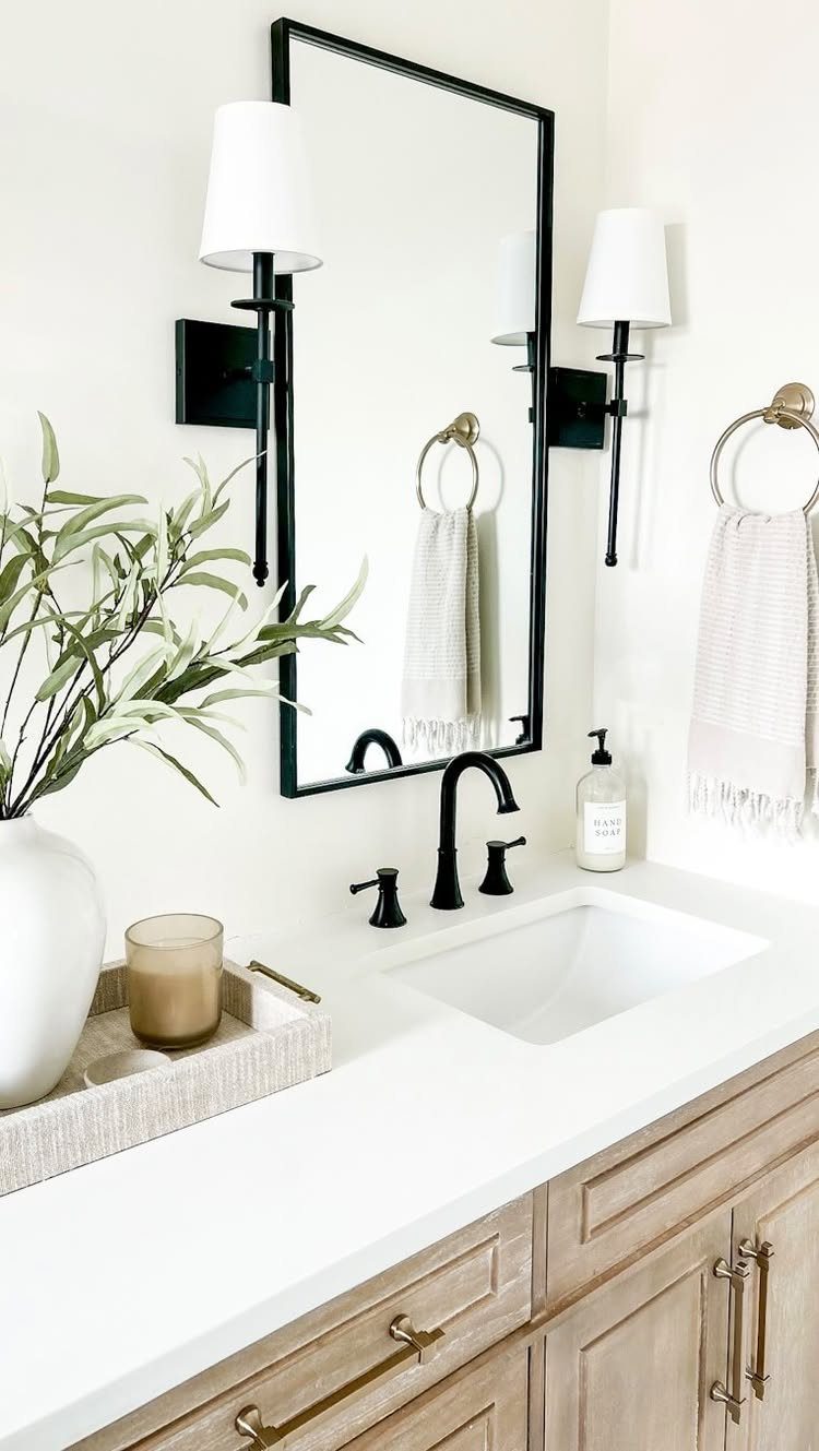 Light wood bathroom vanity with white countertop, black-framed mirror, and white sconces.