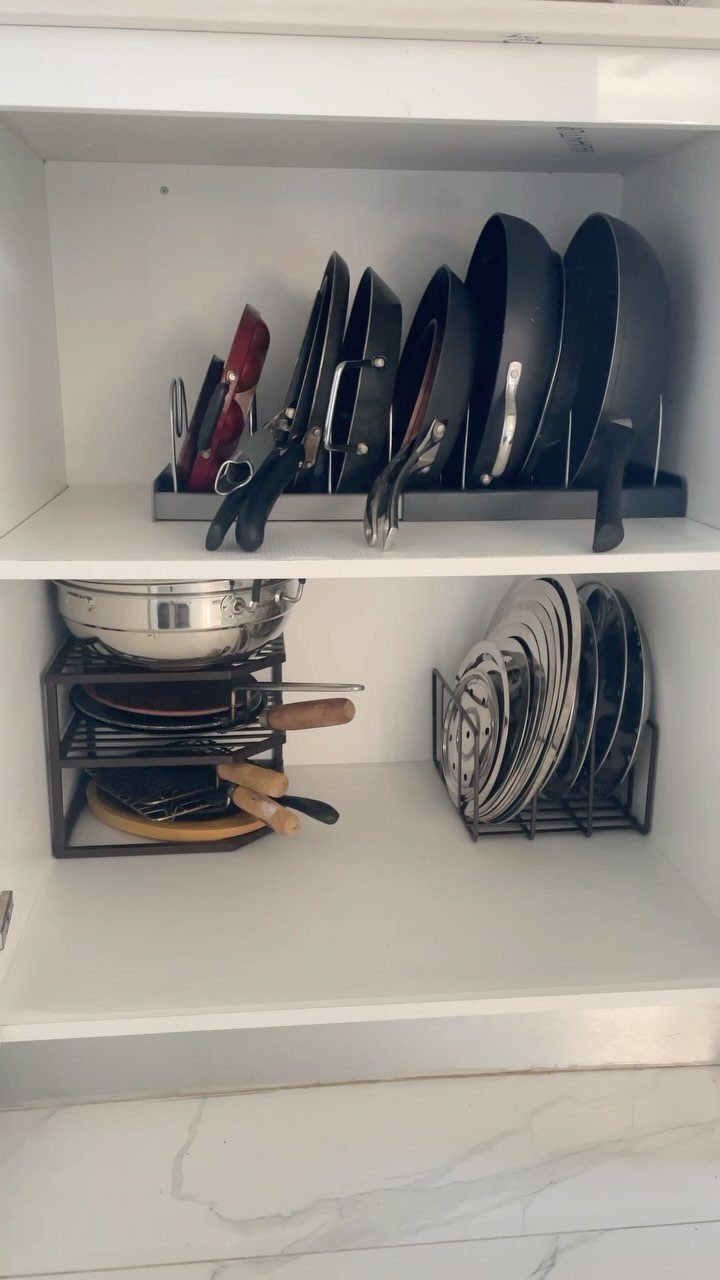 Pan lids stored vertically in a black wire rack inside a white cabinet.