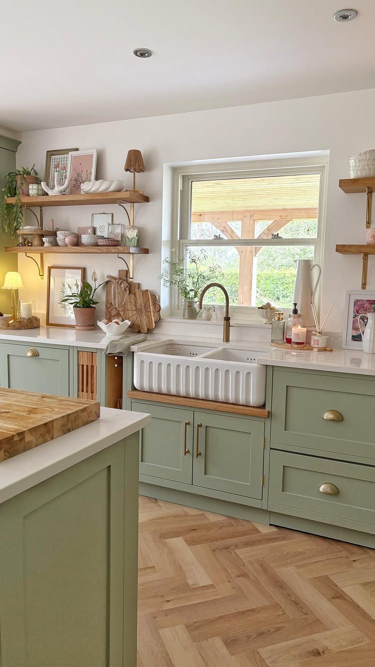 Sage green kitchen with white countertops, herringbone wood floors, and open wooden shelving.